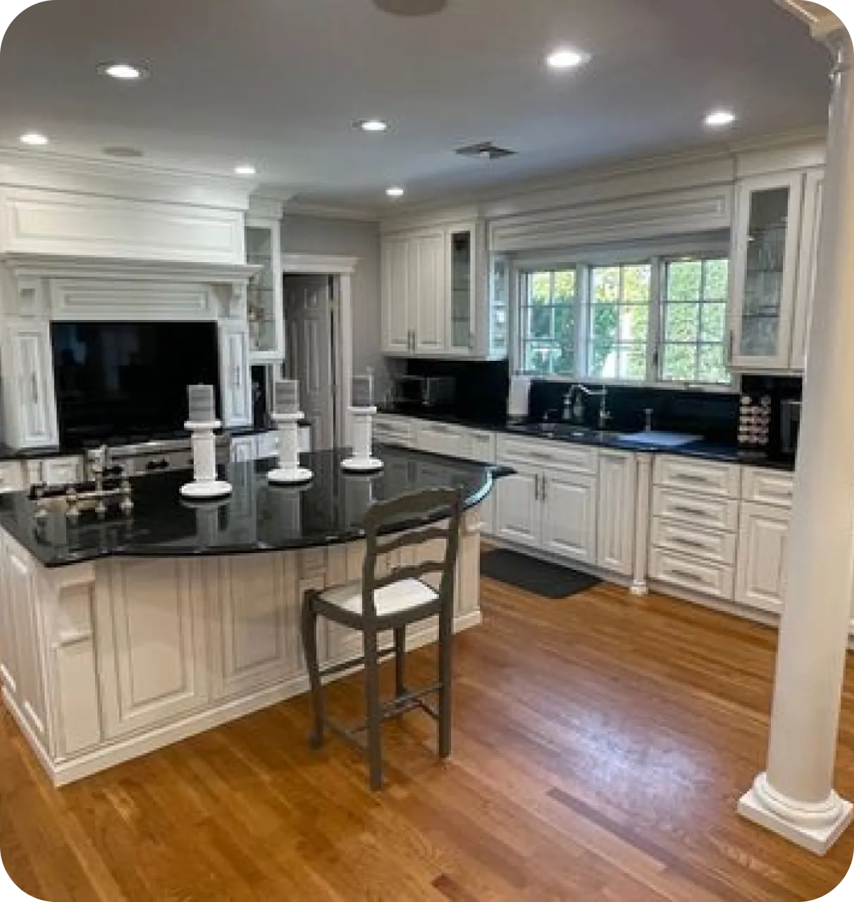 A modern kitchen with white cabinets, a black countertop island with candlesticks, a single chair, and wooden flooring.