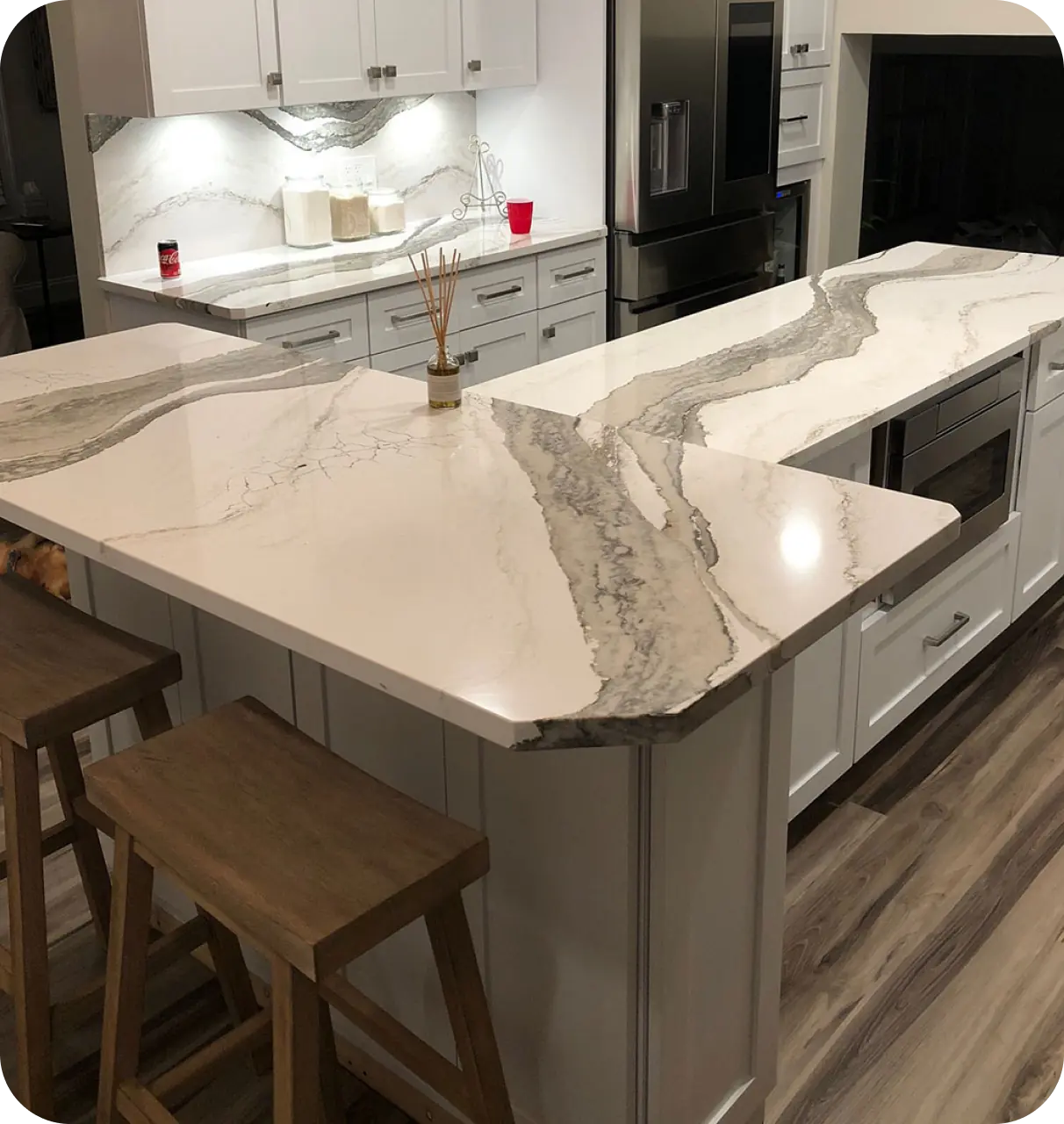 A modern kitchen with a marble countertop island, white cabinets, and wooden stools. A stove and oven are built into the island. The floor features wood-grain-style tiles.