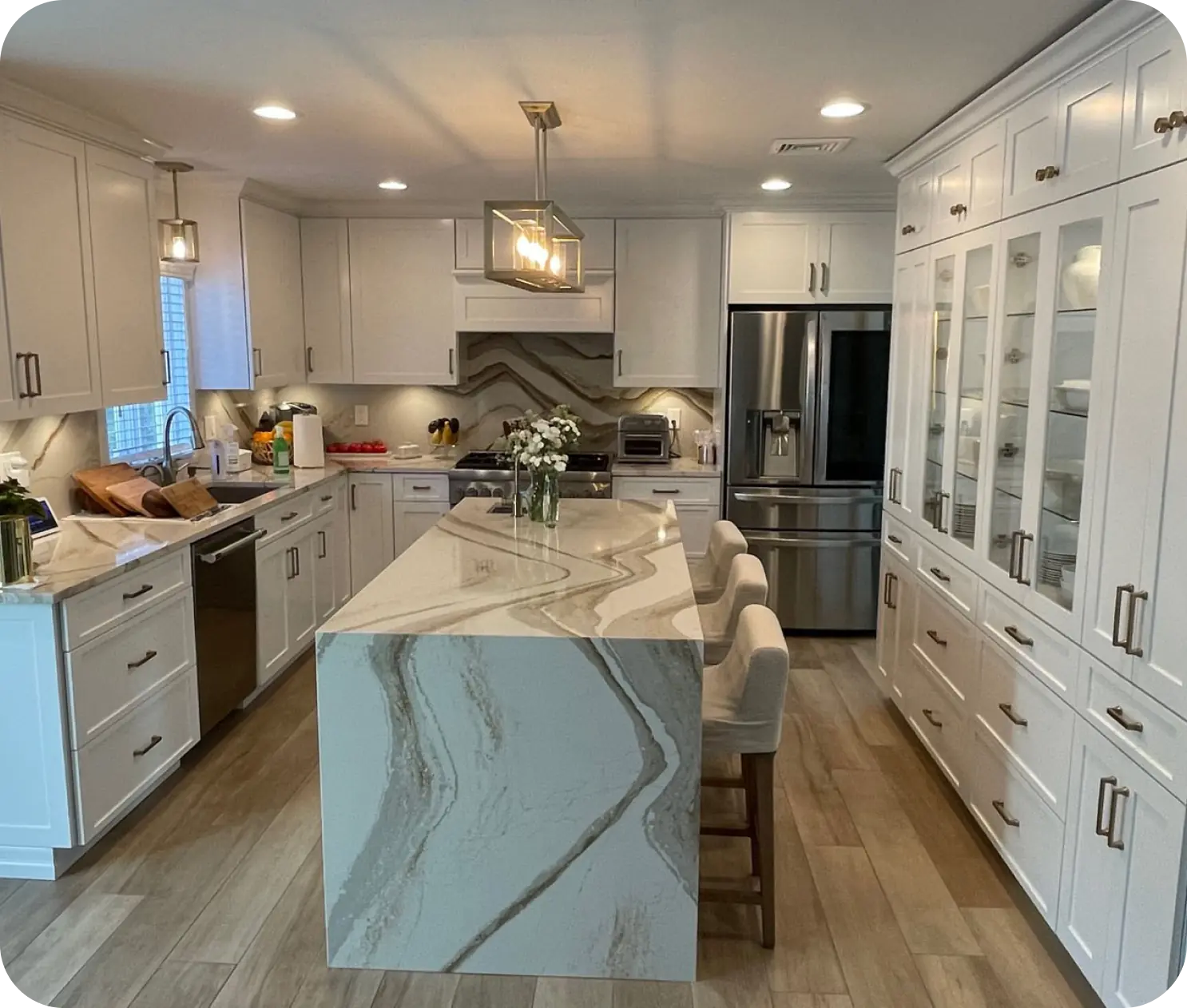 Modern kitchen with a marble island, white cabinets, stainless steel appliances, and hardwood floors. Three upholstered stools are at the island, with pendant lighting above.