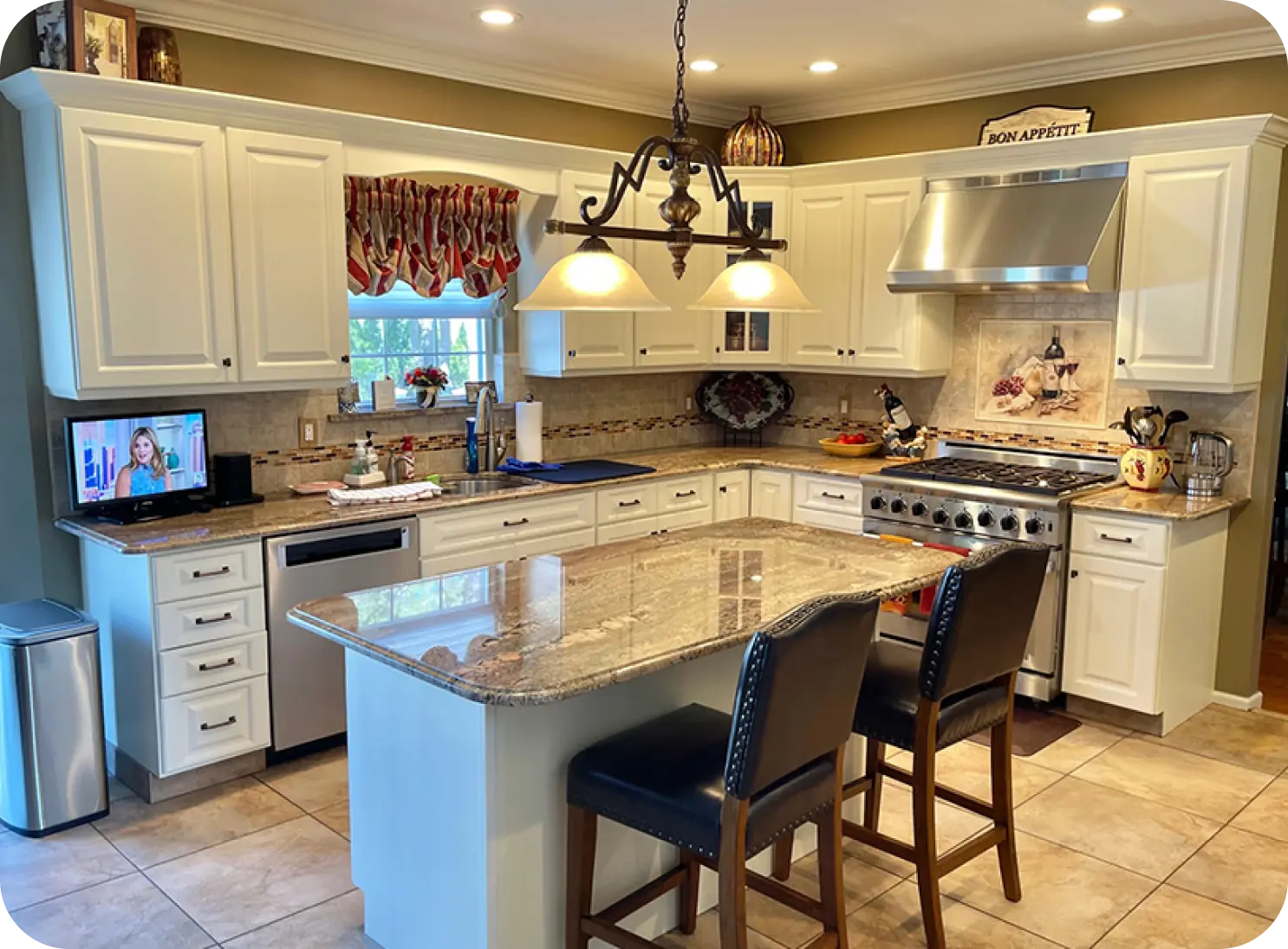 A modern kitchen with white cabinets, granite countertops, a stainless steel stove, and a small TV on the counter. Two chairs are placed at the island. Overhead lights illuminate the space.