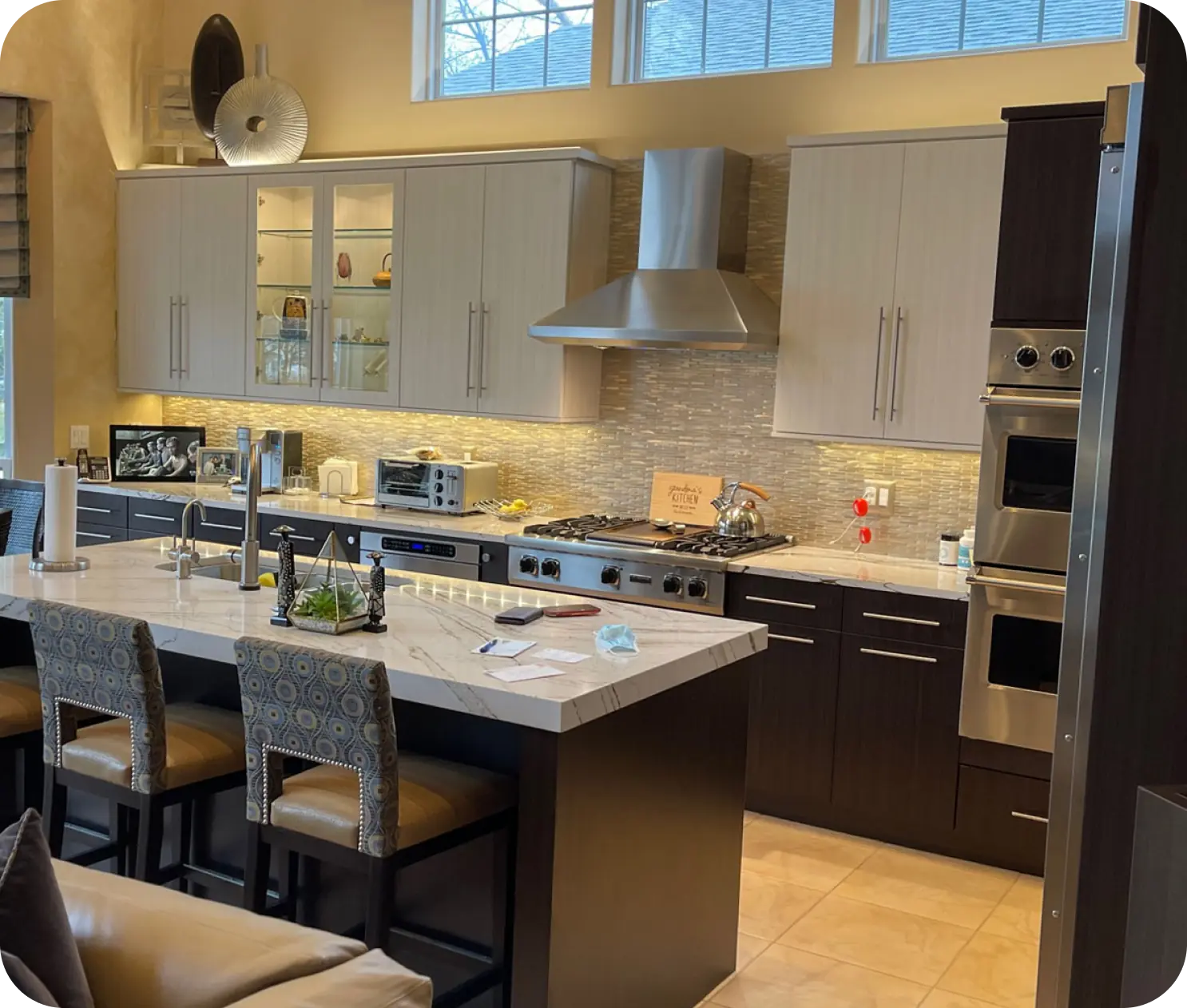 Modern kitchen with dark cabinets, stainless steel appliances, and a marble island with three chairs. Light filters through upper windows, highlighting the backsplash and countertop items.