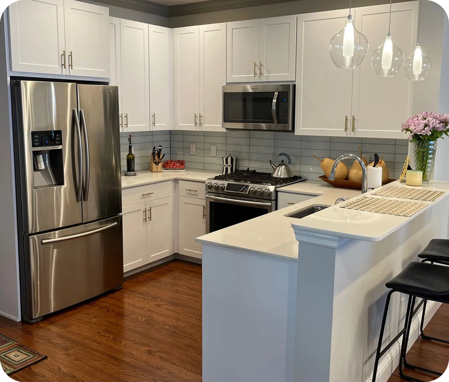 Modern kitchen with white cabinets, stainless steel appliances, and a small island with two stools. Three pendant lights hang above the island. A bouquet of flowers is on the counter.