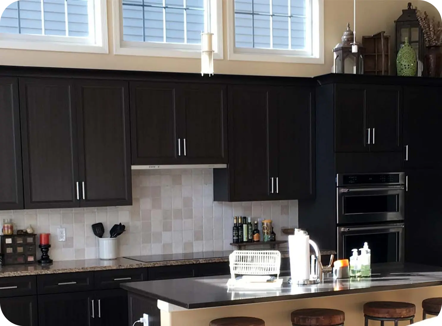 Modern kitchen with dark wood cabinets, stainless steel appliances, and a central island with bar stools. Bright natural light enters through large windows above.