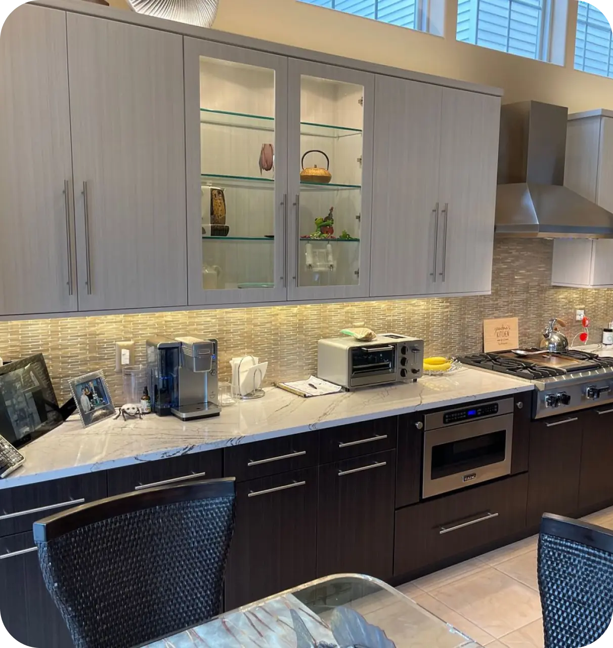 Modern kitchen with dark lower cabinets, light upper cabinets, stainless steel appliances, a coffee maker, and toaster oven on a marble countertop. Backsplash features light brown tiles.