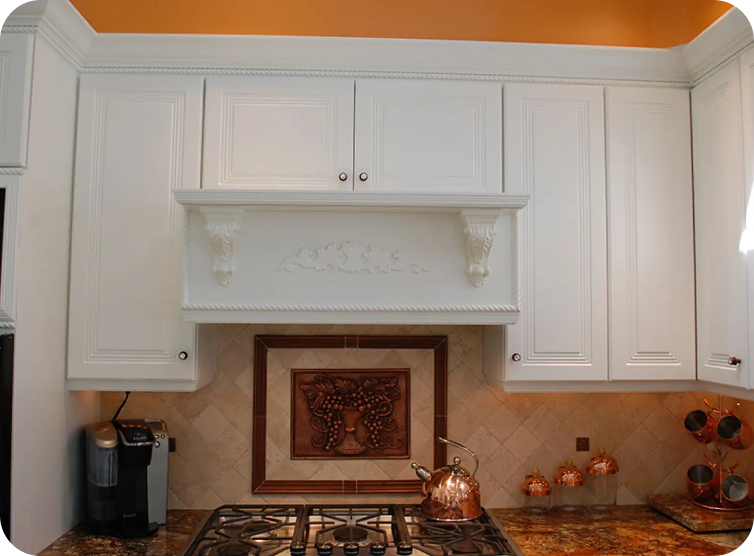 A white kitchen cabinet set with decorative molding above a stovetop. A detailed backsplash features a fruit design. Copper kettles and a holder are on the granite counter.