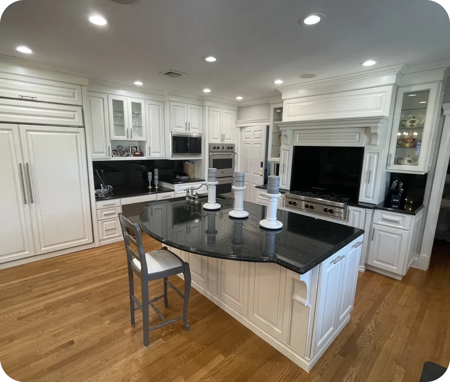 Spacious kitchen with white cabinetry, a large black granite island, and wooden flooring. Features stainless steel appliances, chairs, and decorative candlesticks on the island.