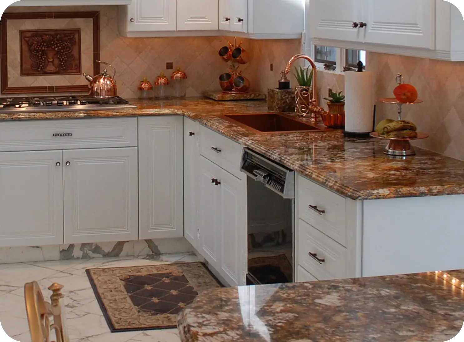 A modern kitchen with white cabinets, marble countertops, a copper sink, and various decor. There's a gas stove on the left and a dishwasher below the counter. A rug is on the tiled floor.